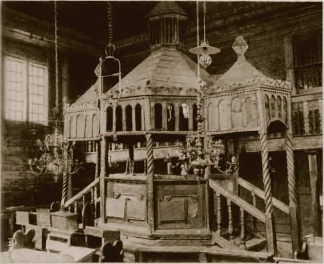 Sepia interior photograph of the domed wooden bimah with arcades and stairways, hanging lamps, and plank walls with windows behind.