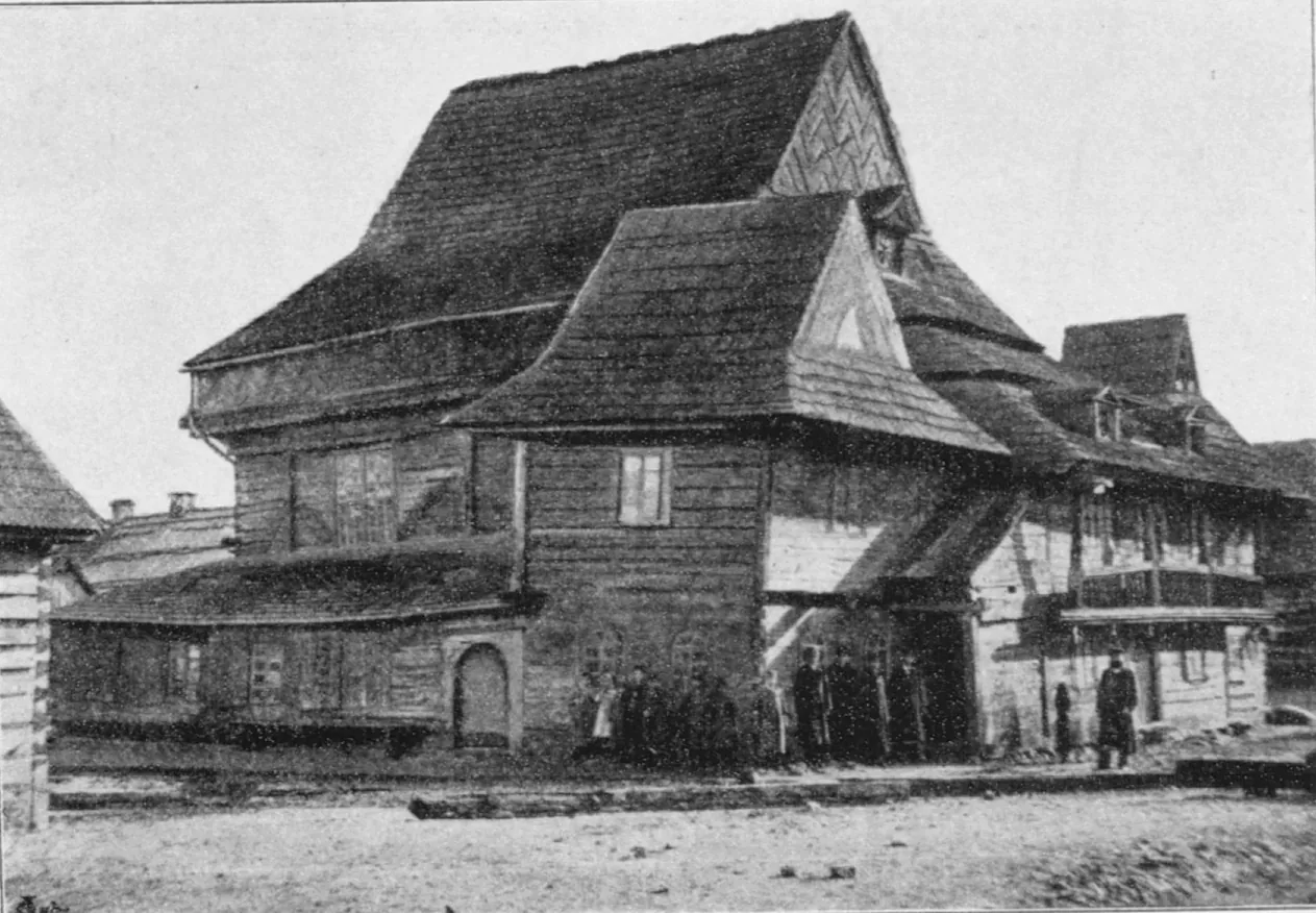 Black-and-white photograph of the timber synagogue from a corner, with steep overlapping roofs, a two-storey annex and people standing along the wall.