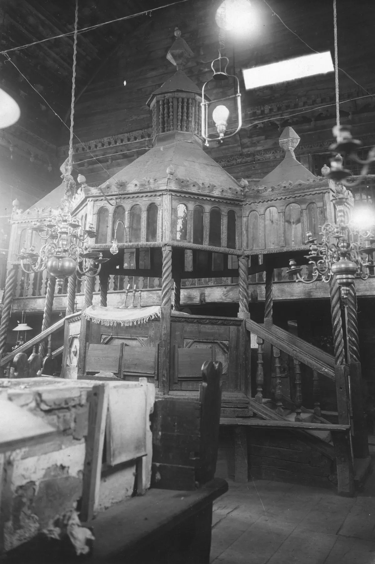 Black-and-white interior photograph of an ornate wooden bimah pavilion with arcaded upper storey and domed roof, stairs on both sides, and brass chandeliers.