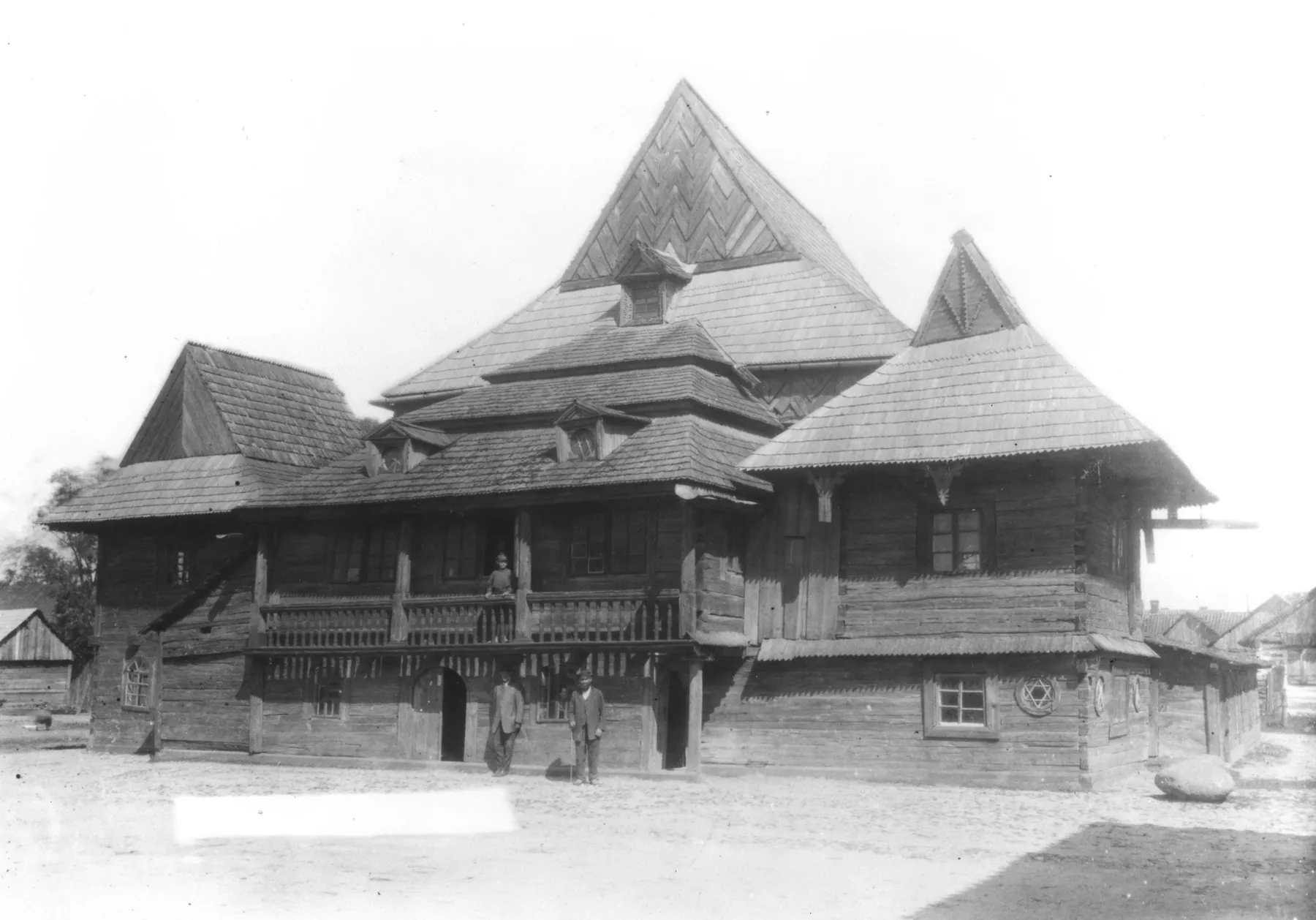 Black-and-white photograph of a large timber synagogue with a steep central roof and several smaller pyramid-roofed pavilions and galleried annexes around it, a few people standing in front.